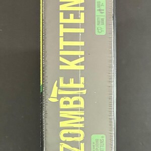 May include: A sealed box of the party game "Zombie Kittens" by Exploding Kittens. The box is dark green with yellow text, including the game title and age/player information. The game is for ages 7+ and 2-5 players.