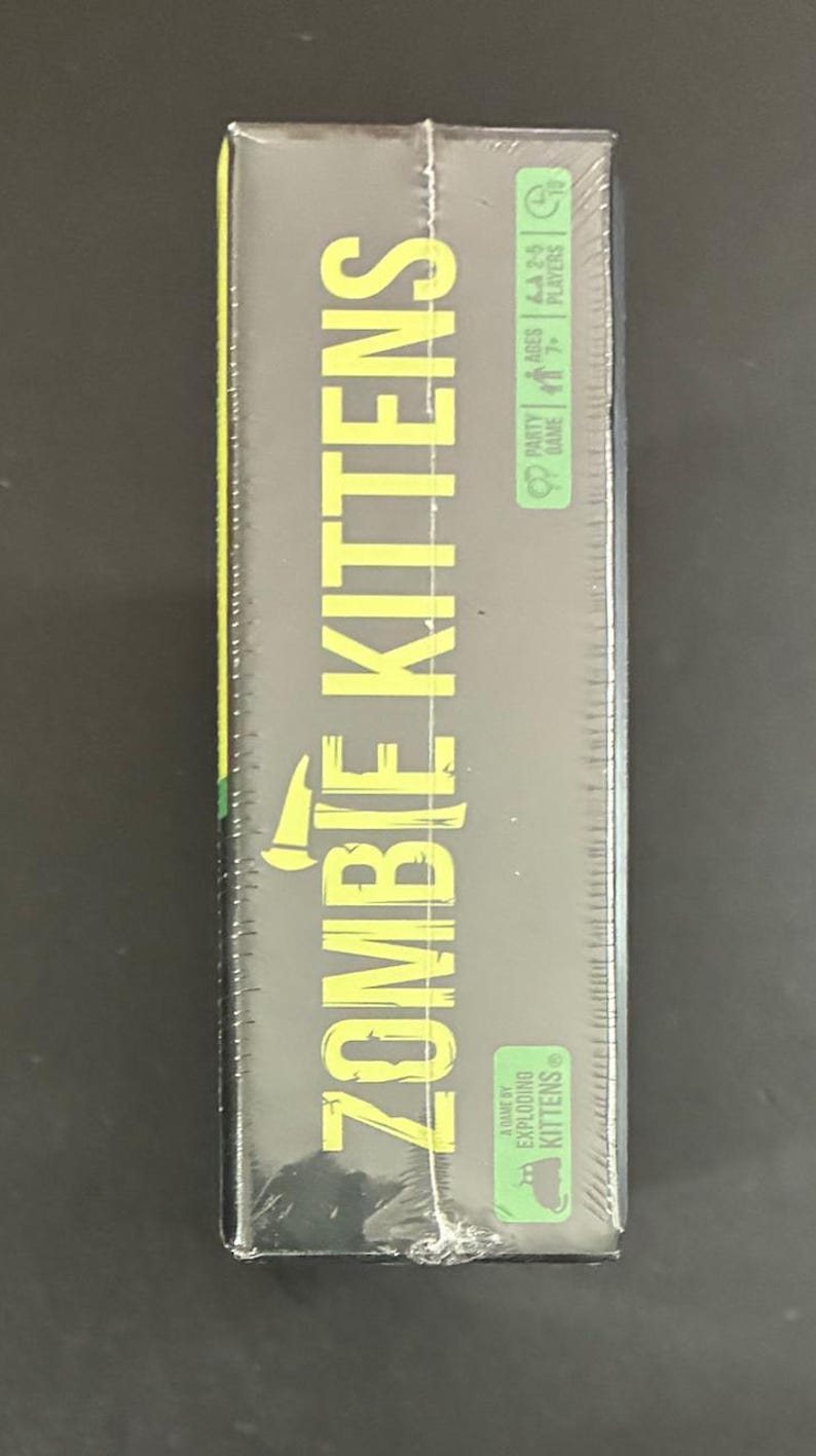 May include: A sealed box of the party game "Zombie Kittens" by Exploding Kittens. The box is dark green with yellow text, including the game title and age/player information. The game is for ages 7+ and 2-5 players.