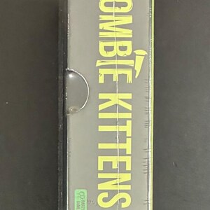 May include: A sealed box for the card game "Zombie Kittens." The game title is in large, yellow-green letters. The box is black with green accents and includes the "Exploding Kittens" logo.