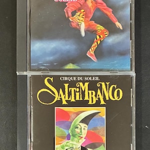 May include: Two CD cases featuring Cirque du Soleil performances. The top case displays "Cirque Du Soleil" with an image of a performer in a colorful costume. The bottom case reads "Cirque Du Soleil Saltimbanco" with a green-faced character.