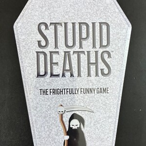 May include: A coffin-shaped game box with the title "STUPID DEATHS" in large, bold letters. The box features a textured, light gray background and an illustration of the Grim Reaper. The text "THE FRIGHTFULLY FUNNY GAME" is also visible.