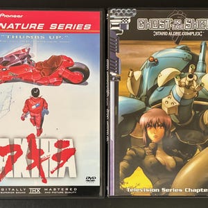 May include: Two DVD cases featuring anime artwork. The left case displays the title AKIRA with a red motorcycle and a character in a red jacket. The right case shows the title GHOST IN THE SHELL STAND ALONE COMPLEX with a blue robotic figure and a character.