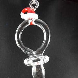 May include: A clear glass ornament shaped like a baby's pacifier with a red and white Santa hat on top.
