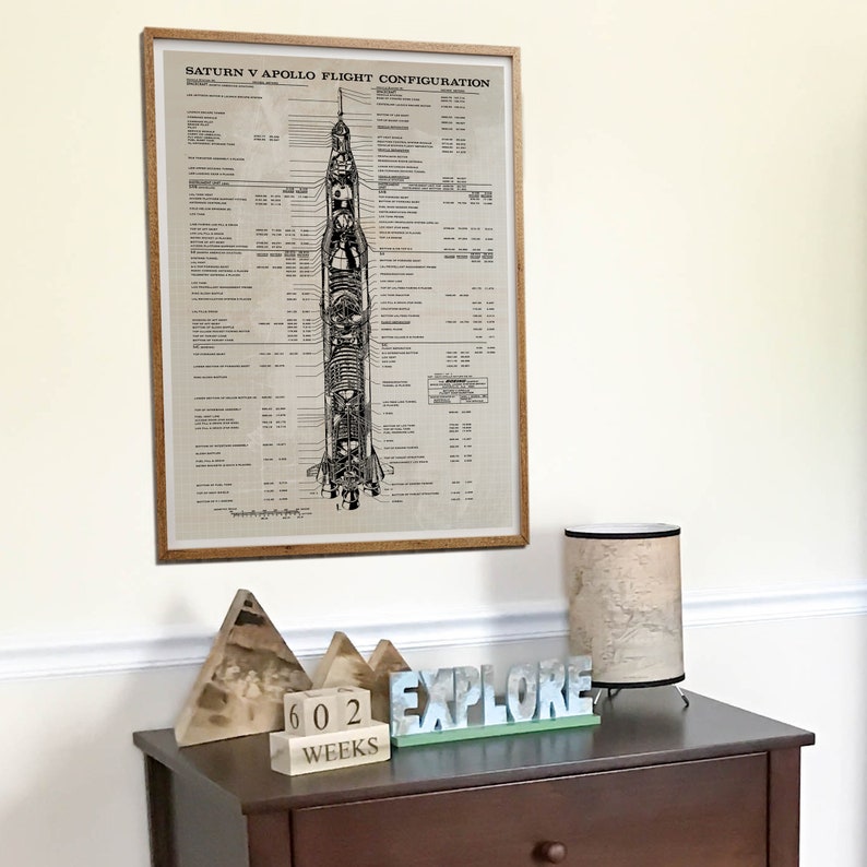 Peut inclure: Une illustration en noir et blanc encadr&eacute;e d'une fus&eacute;e Saturn V avec le texte "Saturn V Apollo Flight Configuration". L'illustration est sur un fond beige. Le cadre est en bois brun. L'image est sur un mur blanc au-dessus d'une commode brune avec une lampe, des blocs de bois qui disent "602 Weeks", et une pancarte en bois qui dit "Explore".