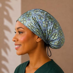 May include: A light blue scrub cap featuring a repeating pattern of green four-leaf clovers and small purple diamond shapes. The cap has a rounded shape with a black elastic drawstring cord at the back for an adjustable fit, covering the head.