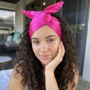 May include: A vibrant pink satin headband with a knotted design and bow detail. The headband is worn on the head and is a striking accessory. The hair is dark and curly.