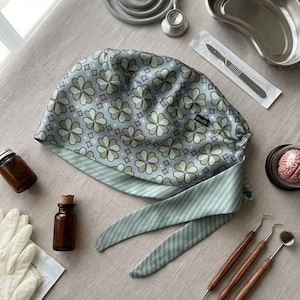 May include: A light blue medical scrub cap with a clover pattern, alongside medical tools and supplies. The cap features a clover pattern in shades of green and purple. Other items include a stethoscope, surgical tools, and a kidney dish.