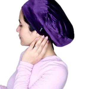 May include: A deep purple satin bonnet. The bonnet is smooth and shiny, with a rounded shape that covers the head. The bonnet is designed to protect hair while sleeping. The brand name "Bliss Silk" is visible on the side.