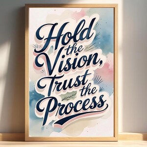 Op de afbeelding: Een ingelijste kunstdruk met de woorden "Hold the Vision, Trust the Process" in een gestileerd schrift. Het kunstwerk heeft een aquarel achtergrond met blauwe, roze en witte tinten. De lijst is lichtbruin.