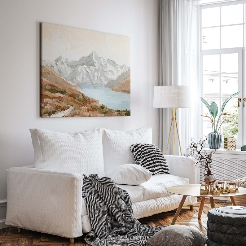 Op de afbeelding: Een witte bank met een zwart-wit gestreept kleed en een kussen. Boven de bank hangt een schilderij van een bergketen met een meer op de voorgrond. Naast de bank staat een gouden staande lamp met een witte kap.