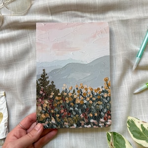 Può includere: Un taccuino rettangolare con un dipinto a impasto materico sulla copertina. L'opera raffigura un paesaggio con un cielo rosa pallido, dolci colline grigie e un primo piano ricco di fogliame denso nei toni del verde, dell'oro e del ruggine.