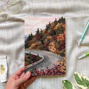 May include: An oil painting on a rectangular canvas depicting a winding gray road curving through a dense forest with autumn foliage in shades of orange, red, brown, and dark green under a pale, soft pink sky with visible thick paint texture.