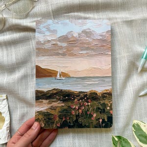 May include: A rectangular notebook featuring a textured impasto oil painting on the cover. The artwork depicts a coastal scene with a sailboat on calm water, a rocky shoreline with small pink flowers in the foreground, and a soft, cloudy sky.