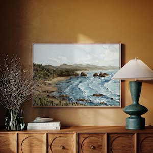 K&ouml;nnte beinhalten: Ein gerahmtes Landschaftsgem&auml;lde zeigt eine K&uuml;stenlandschaft mit Sandstrand, felsiger K&uuml;ste und blauen Meereswellen. Das Kunstwerk wird &uuml;ber einem Holzschrank ausgestellt, neben einer t&uuml;rkisfarbenen Lampe und einer Vase mit getrockneten Zweigen.