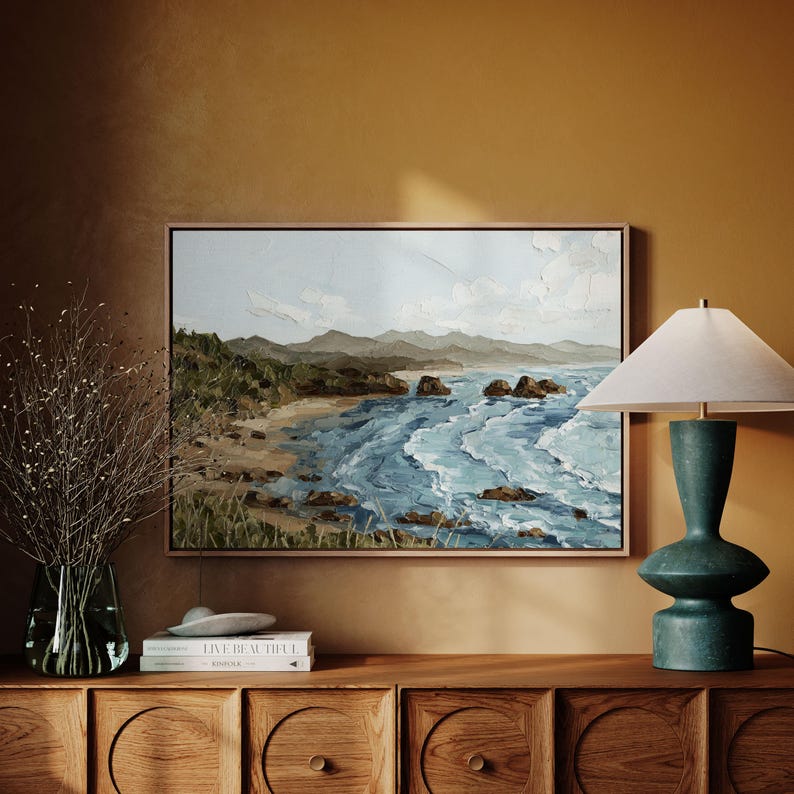 K&ouml;nnte beinhalten: Ein gerahmtes Landschaftsgem&auml;lde zeigt eine K&uuml;stenlandschaft mit Sandstrand, felsiger K&uuml;ste und blauen Meereswellen. Das Kunstwerk wird &uuml;ber einem Holzschrank ausgestellt, neben einer t&uuml;rkisfarbenen Lampe und einer Vase mit getrockneten Zweigen.