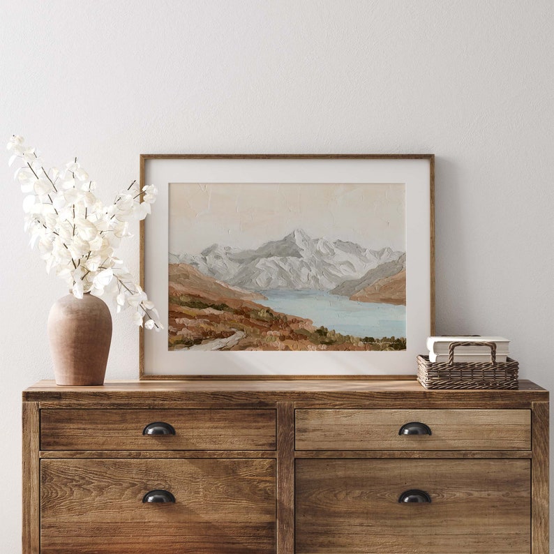 Op de afbeelding: Een ingelijst schilderij van een bergketen met een meer op de voorgrond. De bergen zijn met sneeuw bedekt en het meer is lichtblauw. Het schilderij is in een lichtbruine lijst en hangt aan een witte muur.