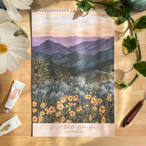 May include: A 2027 wall calendar by Hannah Weisner Art featuring a landscape painting of yellow wildflowers in the foreground, dark green hills in the midground, and purple mountains under a pale peach sky in the background.