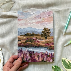 May include: A rectangular notebook featuring a thick, textured oil painting on the cover. The artwork depicts a landscape with a pale pink sky, a single tree, rolling brown hills, and a foreground filled with vibrant purple and pink flowers.