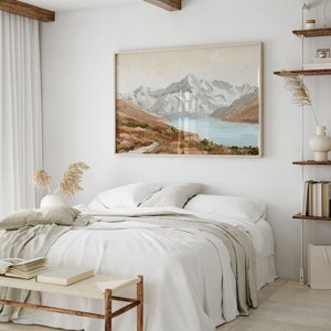 Op de afbeelding: Een wit bed met een beige deken en een wit kussen. Aan de muur achter het bed hangt een ingelijst schilderij van een bergketen met een meer op de voorgrond.