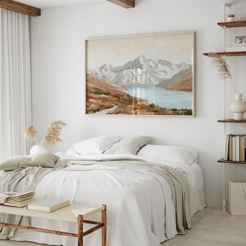 Op de afbeelding: Een wit bed met een beige deken en een wit kussen. Aan de muur achter het bed hangt een ingelijst schilderij van een bergketen met een meer op de voorgrond.