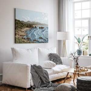 K&ouml;nnte beinhalten: Ein Wohnzimmer mit wei&szlig;em Sofa, Kissen und einer gestreiften Decke. &Uuml;ber dem Sofa h&auml;ngt ein quadratisches &Ouml;lgem&auml;lde, das eine K&uuml;stenlandschaft mit blauem Wasser, einem Sandstrand und gr&uuml;nen H&uuml;geln zeigt.