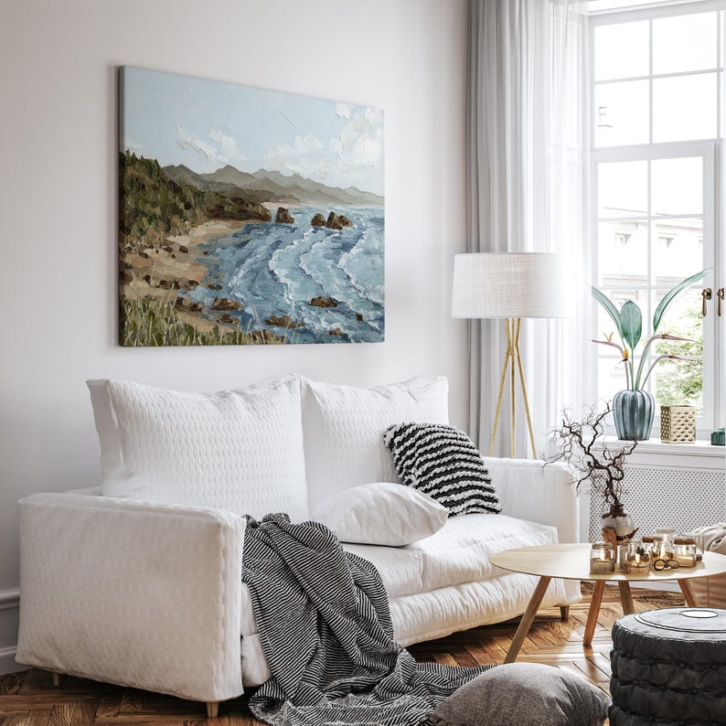 K&ouml;nnte beinhalten: Ein Wohnzimmer mit wei&szlig;em Sofa, Kissen und einer gestreiften Decke. &Uuml;ber dem Sofa h&auml;ngt ein quadratisches &Ouml;lgem&auml;lde, das eine K&uuml;stenlandschaft mit blauem Wasser, einem Sandstrand und gr&uuml;nen H&uuml;geln zeigt.