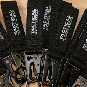 May include: A group of black keychains with silver metal clips and rings. Each keychain has a black fabric strap with the text "TACTICAL GEARLINES" printed in white.