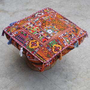 Puede incluir: Un cojín de suelo cuadrado colorido con un diseño de patchwork y detalles bordados. El cojín está hecho de retazos de tela cosidos juntos y tiene borlas en los bordes.