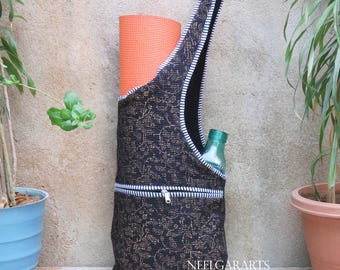 May include: A black yoga mat bag with a gold floral pattern and a white zipper. The bag has a black strap and holds an orange yoga mat and a green water bottle. The bag is designed to carry yoga essentials.