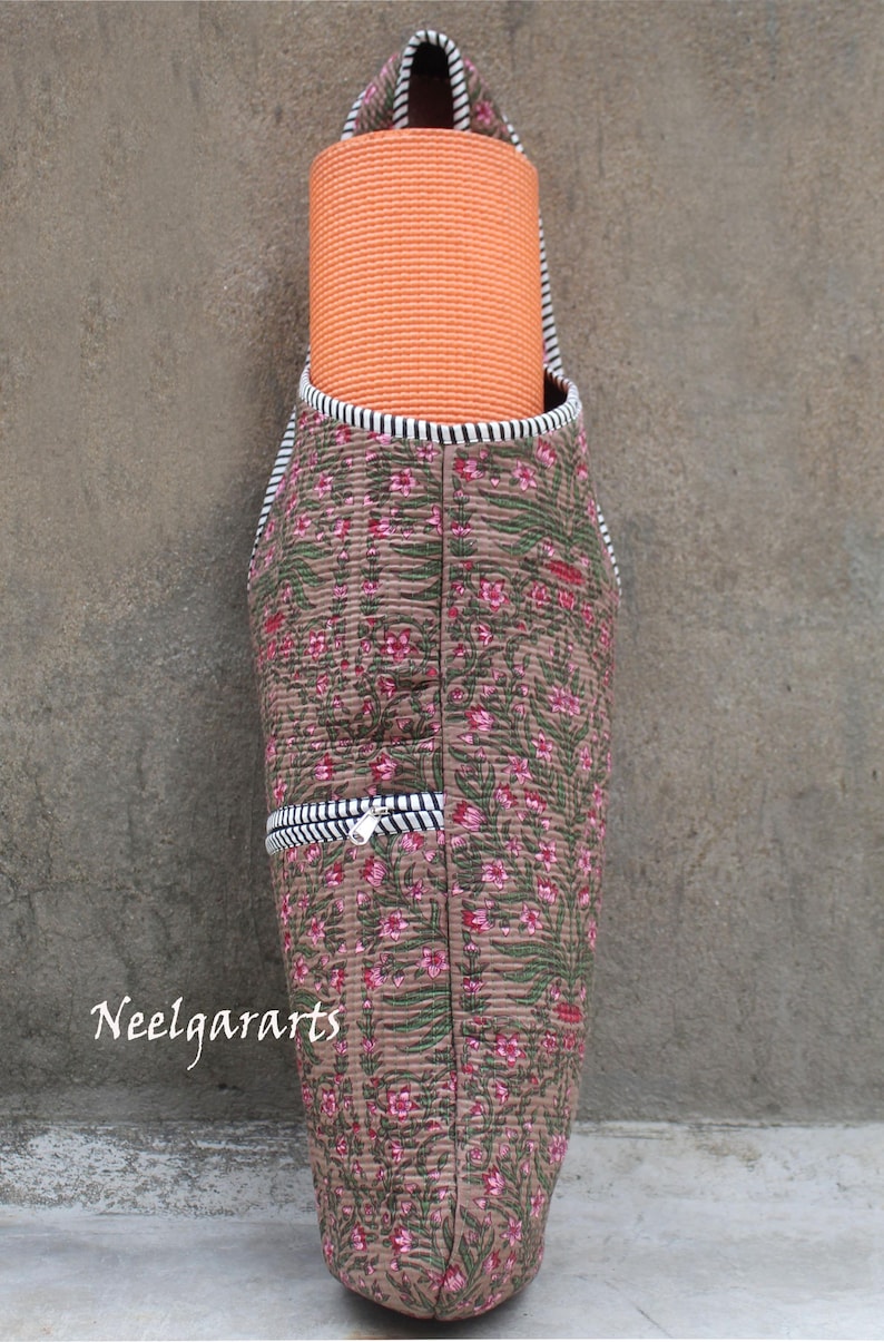 K&ouml;nnte beinhalten: Gesteppte Yogamatten-Tasche mit floralem Muster in Braun, Gr&uuml;n und Rosa. Die Tasche hat eine schwarz-wei&szlig; gestreifte Bord&uuml;re und eine Rei&szlig;verschlusstasche. Eine orangefarbene Yogamatte befindet sich im Inneren. Der Text "Neelgararts" ist sichtbar.