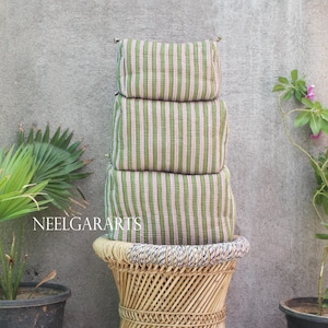 May include: Three green and white striped cushions stacked on a woven stool. The cushions have a vertical stripe pattern. The stool is made of light-coloured woven material. The background is a grey wall with plants on either side.