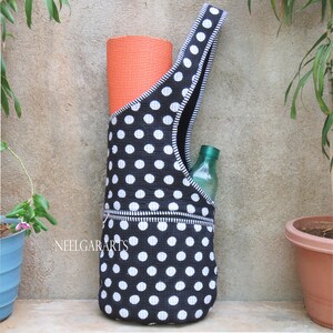 May include: A black yoga mat bag with white polka dots, holding an orange yoga mat and a green water bottle. The bag has a zippered pocket and a black and white striped strap. The text "NEELGARARTS" is visible on the bag.
