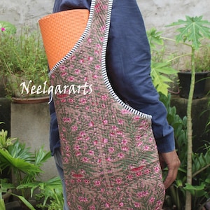 May include: A brown yoga mat bag with a floral pattern in pink and green, carried over the shoulder. The bag has a striped trim and holds an orange yoga mat. The text "Neelgararts" is visible on the bag.