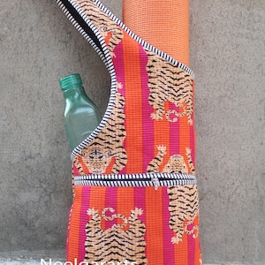 Könnte beinhalten: Eine Yogamatten-Tragetasche mit Tigerdruck. Die Tasche hat orange und pinkfarbene vertikale Streifen, eine schwarz-weiß gestreifte Bordüre und eine Reißverschlusstasche. Eine orangefarbene Yogamatte und eine grüne Flasche sind sichtbar.