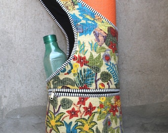May include: A yoga mat carrier with a colorful floral pattern, holding an orange yoga mat and a green water bottle. The carrier has a black zipper and a black and white striped strap. The text "Neelgararts" is visible.