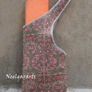 K&ouml;nnte beinhalten: Eine braune Yogamatten-Tragetasche mit einem floralen Muster in Rosa und Gr&uuml;n. Die Tasche hat einen Riemen mit schwarz-wei&szlig;en Streifen und h&auml;lt eine orangefarbene Yogamatte. Der Text "Neelgararts" ist sichtbar.