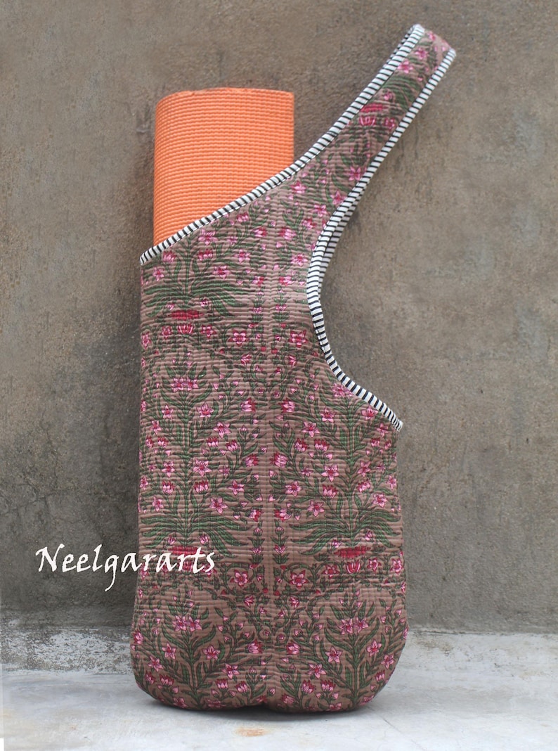 K&ouml;nnte beinhalten: Eine braune Yogamatten-Tragetasche mit einem floralen Muster in Rosa und Gr&uuml;n. Die Tasche hat einen Riemen mit schwarz-wei&szlig;en Streifen und h&auml;lt eine orangefarbene Yogamatte. Der Text "Neelgararts" ist sichtbar.