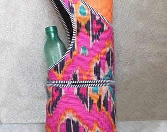 May include: A pink yoga mat bag with a colorful ikat pattern, holding an orange yoga mat and a green glass water bottle. The bag has a black and white striped trim and a shoulder strap.