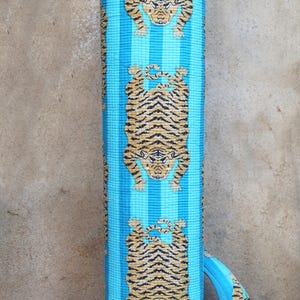 Puede incluir: Una esterilla de yoga a rayas azules y marrones con un patr&oacute;n repetido de tigres. La esterilla tiene una correa de transporte.