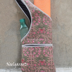 K&ouml;nnte beinhalten: Eine braune Yogamatten-Tasche mit einem rosa-gr&uuml;nen Blumenmuster, die eine orangefarbene Yogamatte und eine gr&uuml;ne Wasserflasche enth&auml;lt. Die Tasche hat ein schwarzes Innenfutter und eine wei&szlig;-schwarz gestreifte Bord&uuml;re. Der Text "Neelgararts" ist sichtbar.