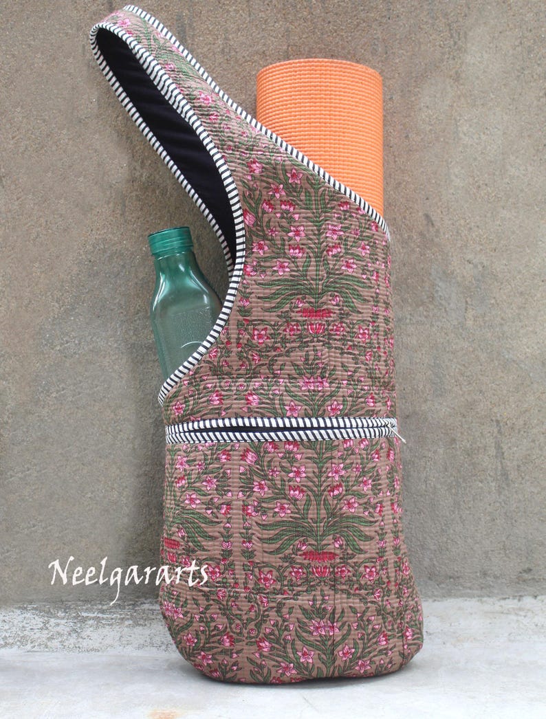 K&ouml;nnte beinhalten: Eine braune Yogamatten-Tasche mit einem rosa-gr&uuml;nen Blumenmuster, die eine orangefarbene Yogamatte und eine gr&uuml;ne Wasserflasche enth&auml;lt. Die Tasche hat ein schwarzes Innenfutter und eine wei&szlig;-schwarz gestreifte Bord&uuml;re. Der Text "Neelgararts" ist sichtbar.