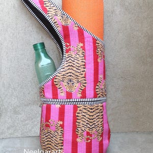 Puede incluir: Una esterilla de yoga y una botella de agua se guardan en una bolsa de yoga a rayas rosas y rojas con estampado de tigre. La bolsa tiene una cremallera negra y un ribete a rayas blancas y negras. Una esterilla de yoga naranja está enrollada dentro.