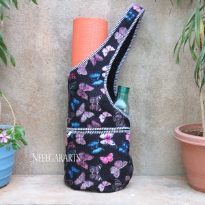 Peut inclure: Sac de tapis de yoga noir avec un motif de papillons roses, violets et bleus. Le sac a une sangle noire avec des rayures blanches et une poche zippée. Un tapis de yoga orange et une bouteille d'eau verte sont visibles.