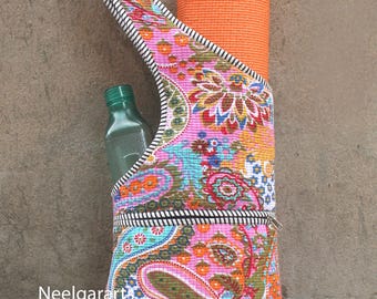 May include: A colorful yoga mat bag with a paisley pattern in pink, blue, and orange. The bag has a zipper closure and a strap with a black and white striped design. An orange yoga mat and a green water bottle are inside.