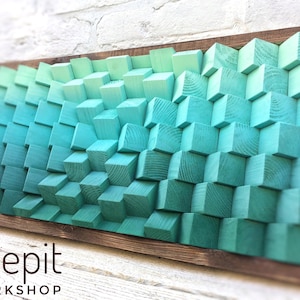May include: A framed wall art piece made of wood blocks painted in shades of teal and green. The blocks are arranged in a 3D pattern, creating a wave-like effect. The frame is made of dark wood.