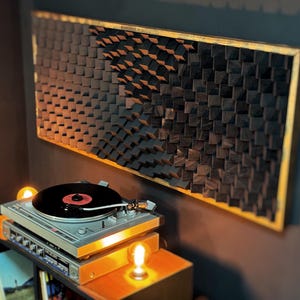 Op de afbeelding: Een zwart en gouden omlijst akoestisch paneel gemaakt van houten blokken. Het paneel is gemonteerd op een muur naast een platenspeler.