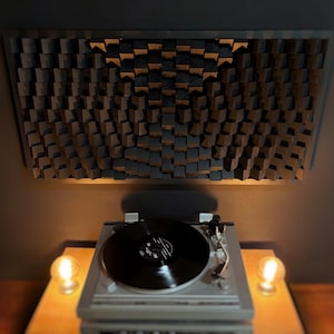 May include: A turntable with a black vinyl record sits on a wooden surface, illuminated by two warm light bulbs. Above, a dark wooden acoustic panel with a geometric design is mounted on the wall, creating a modern audio setup.