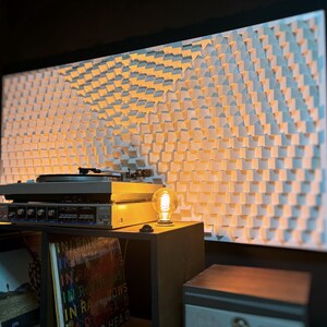 May include: A home audio setup featuring a turntable, amplifier, and a decorative wall panel. The wall panel has a repeating pattern of white, square blocks. A vintage-style light bulb sits on a small table next to the audio equipment.