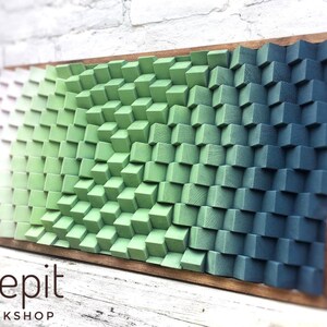 May include: A wall art piece made of wood blocks painted in shades of green and blue. The blocks are arranged in a wave pattern, creating a 3D effect. The piece is framed in a natural wood frame. The text "shepit WORKSHOP" is visible at the bottom of the image.