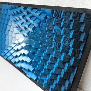 Op de afbeelding: Een blauw en wit abstract wandkunstwerk gemaakt van houten blokken. De blokken zijn gerangschikt in een patroon dat een 3D-effect creëert. Het stuk is ingelijst in zwart.